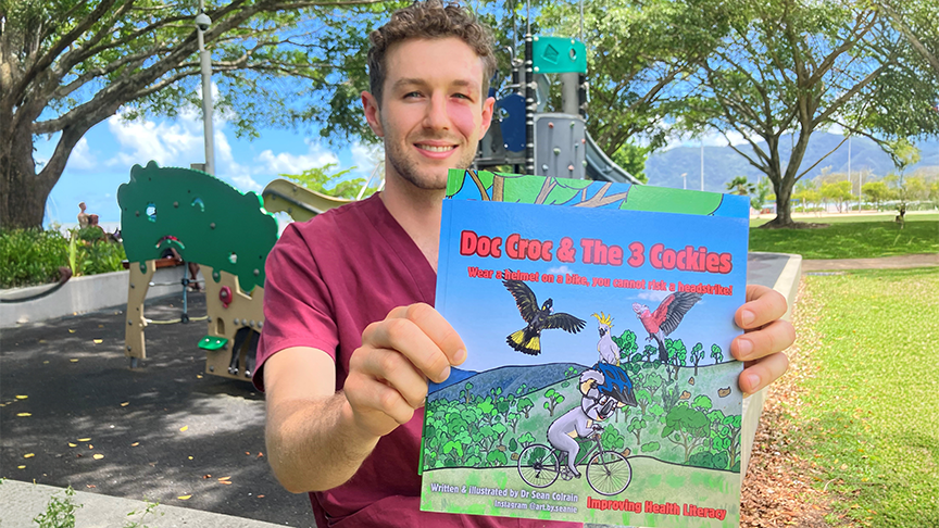Male doctor in scrubs holding a child's picture book out in front of him, standing in front of a playground