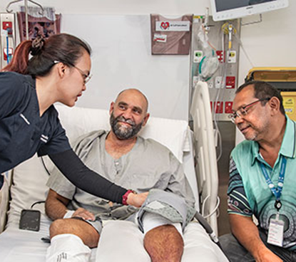 A man lies in a hospital bed, attended by a nurse and a doctor discussing his care.