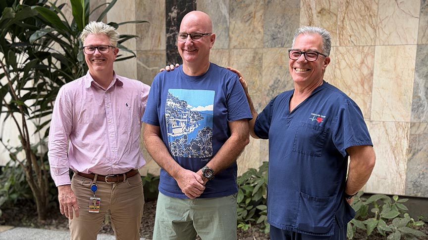Three smiling men, one standing between two others who each have a hand resting on his shoulders