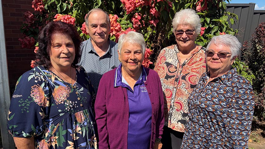 Group of four older women and one man, smiling at the camera in front of a bush covered in flowers