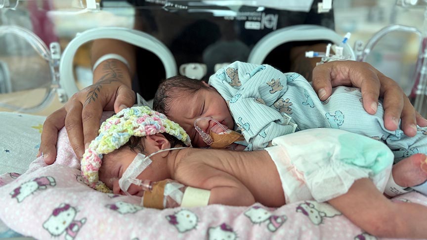 Two very small babies lying next to each other in a hospital incubator with mother's arms coming from outside and laying gently on the babies