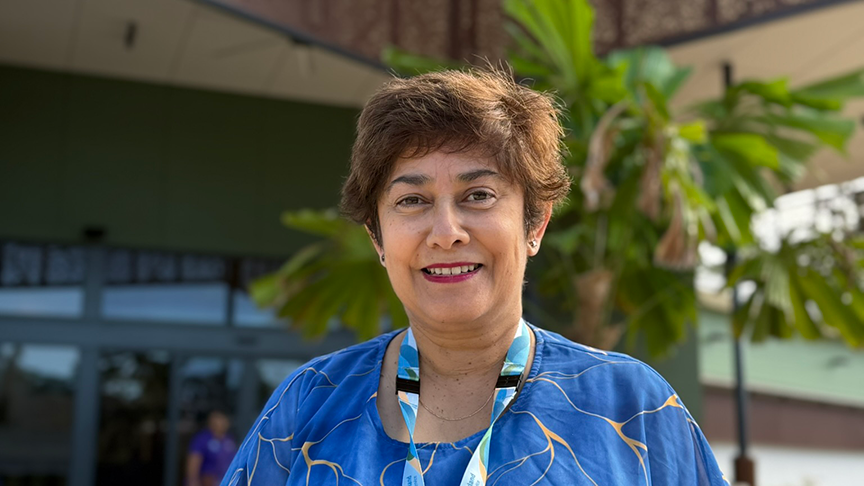 Older brunette woman wearing flowy blue and gold top standing in front of new building and garden smiling at camera