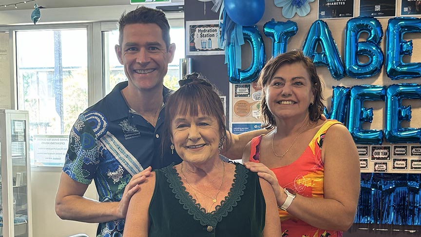 One male and one female clinician standing behind a female consumer in front of a wall with balloons that say Diabetes Week