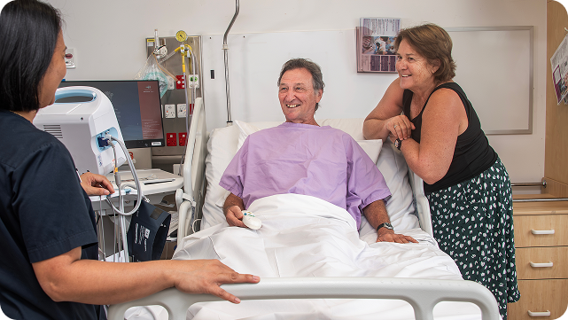 A man lies in a hospital bed, attended by a nurse and a doctor discussing his care.
