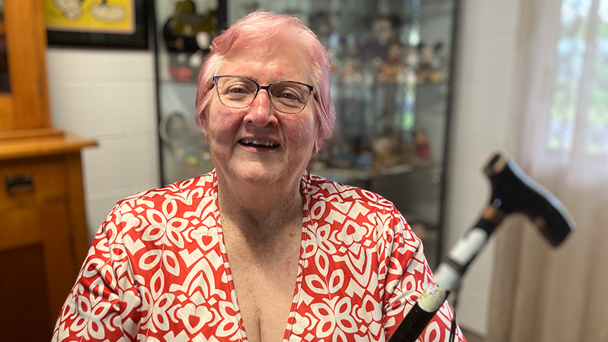 Older woman sitting in her lounge room smiling at the camera holding a walking stick