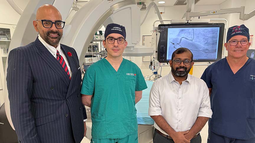 Four doctors standing in a row in front of cardiology medical equipment, two in business clothing and two in scrubs