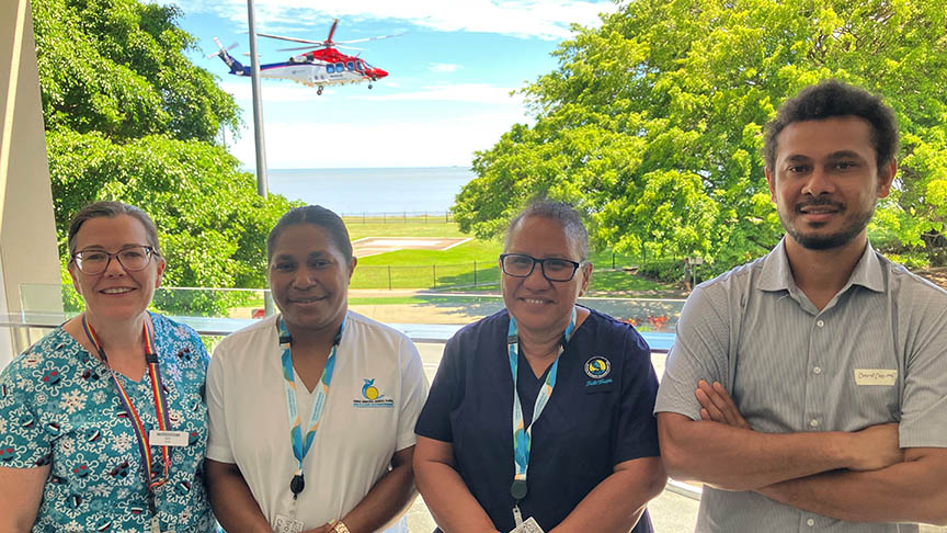 Four clinicians standing in a row on a balcony with helicopter taking off in background