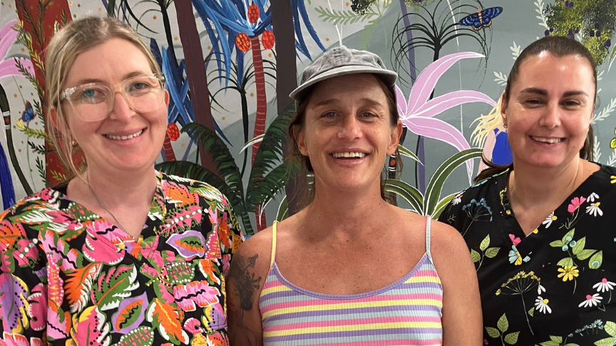 Three women, two in scrubs, standing in front of a rainforest wall mural smiling at the camera
